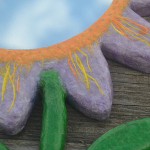 May include: A close-up of a handmade flower-shaped mirror. The mirror has a purple petal design with orange and yellow accents. The stem and leaves are green. The background is a wooden surface.