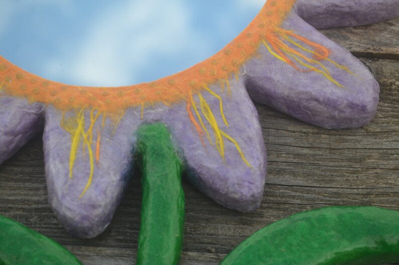 May include: A close-up of a handmade flower-shaped mirror. The mirror has a purple petal design with orange and yellow accents. The stem and leaves are green. The background is a wooden surface.