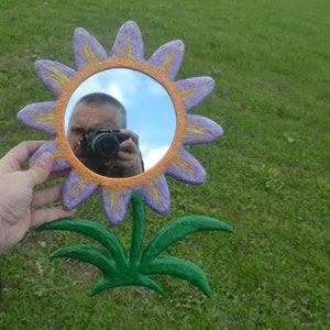 May include: A whimsical, hand-painted flower mirror with a round reflective surface. The flower has purple petals with yellow accents and an orange border. The stem and leaves are green. The mirror is held against a green grassy background.
