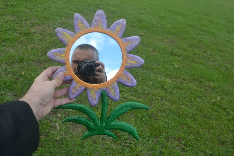 May include: A whimsical, hand-painted flower mirror with a round reflective surface. The flower has purple petals with yellow accents and an orange border. The stem and leaves are green. The mirror is held against a green grassy background.