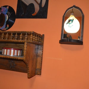 May include: A wooden shelf with a decorative carved trim, two shelves, and a small box on the bottom shelf. The shelf is mounted on a wall painted orange. There is a round mirror with a cat design on the wall above the shelf. A small arched mirror with a hanging light fixture is on the wall to the right of the shelf.