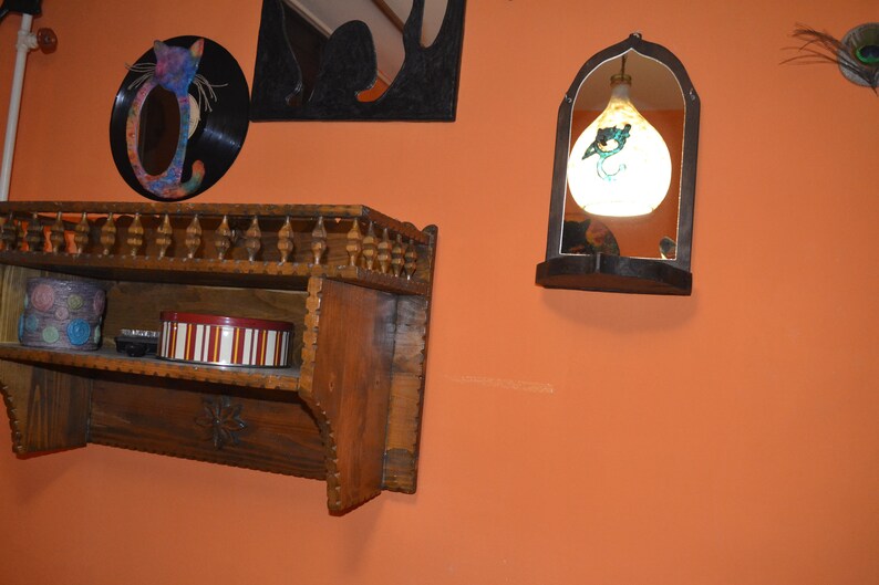 May include: A wooden shelf with a decorative carved trim, two shelves, and a small box on the bottom shelf. The shelf is mounted on a wall painted orange. There is a round mirror with a cat design on the wall above the shelf. A small arched mirror with a hanging light fixture is on the wall to the right of the shelf.