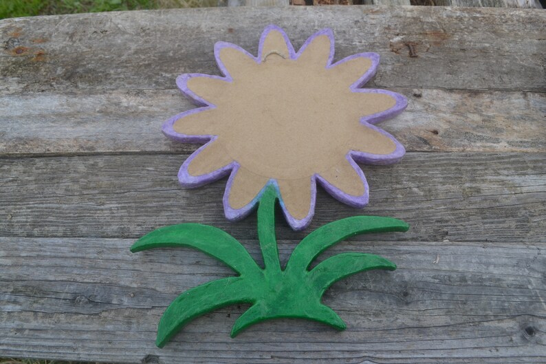 May include: A handmade flower decoration with a light purple outline and a tan center. The flower is separated from the green leaves and stem. The flower and leaves are made of a textured material, possibly paper mache.
