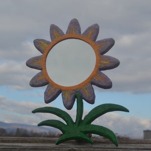 May include: A whimsical, handcrafted mirror shaped like a sunflower. The flower has purple petals with orange and yellow accents, surrounding a circular mirror. The stem and leaves are green, set against a cloudy sky.