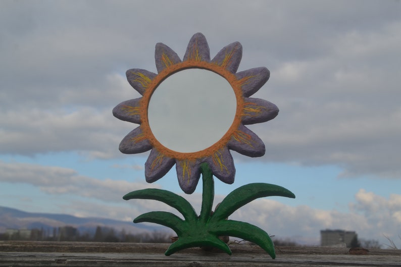 May include: A whimsical, handcrafted mirror shaped like a sunflower. The flower has purple petals with orange and yellow accents, surrounding a circular mirror. The stem and leaves are green, set against a cloudy sky.