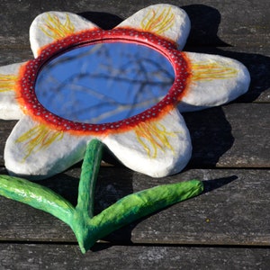 May include: A handcrafted flower-shaped mirror with a white petal design, a red and white dotted border, and a green stem. The mirror reflects a blue sky. The artwork is placed on a weathered wooden surface.