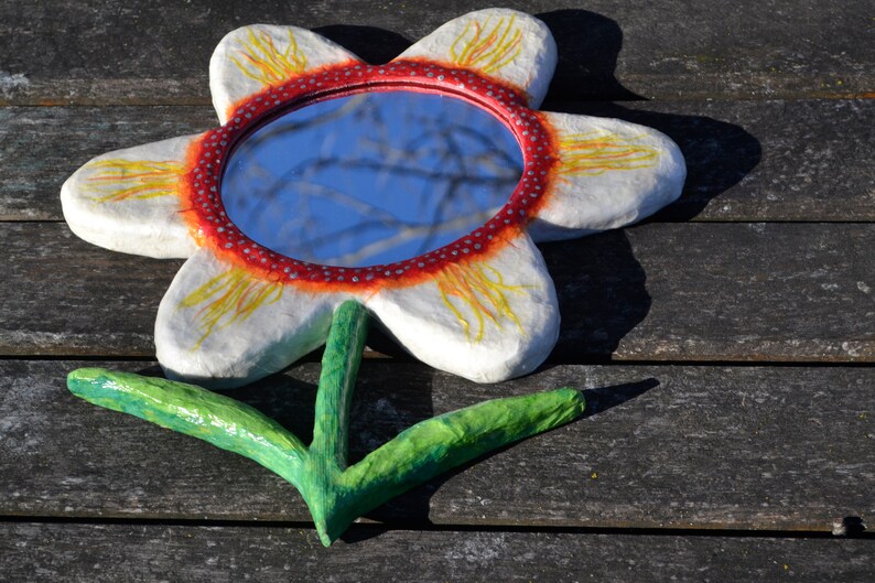 May include: A handcrafted flower-shaped mirror with a white petal design, a red and white dotted border, and a green stem. The mirror reflects a blue sky. The artwork is placed on a weathered wooden surface.