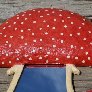 Pu&ograve; includere: Uno specchio a forma di fungo rosso con pois bianchi e un gambo bianco. Lo specchio riflette un cielo blu.