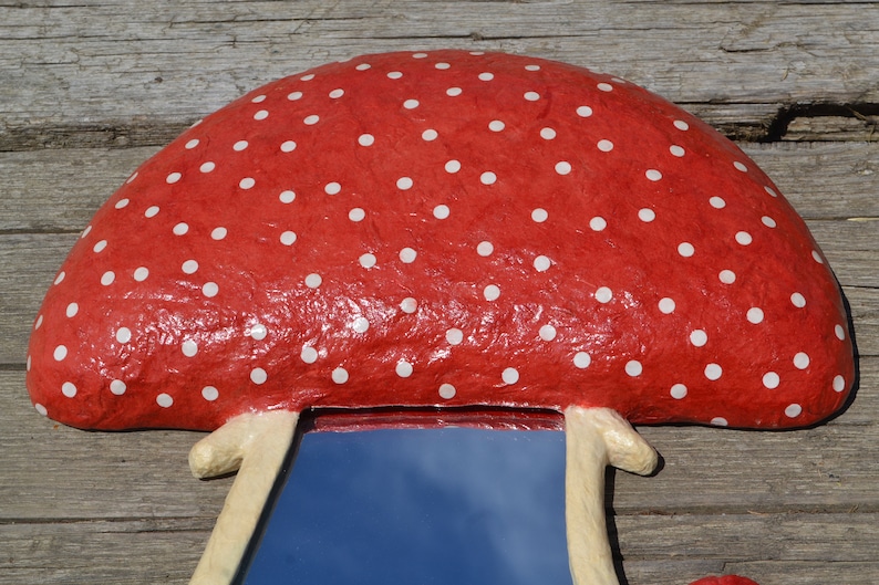 Pu&ograve; includere: Uno specchio a forma di fungo rosso con pois bianchi e un gambo bianco. Lo specchio riflette un cielo blu.