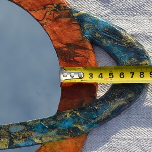 May include: A round mirror with a colorful, textured frame. The frame is painted with orange, blue, and gold accents. The mirror's diameter is approximately 4 inches as measured by a yellow tape measure.