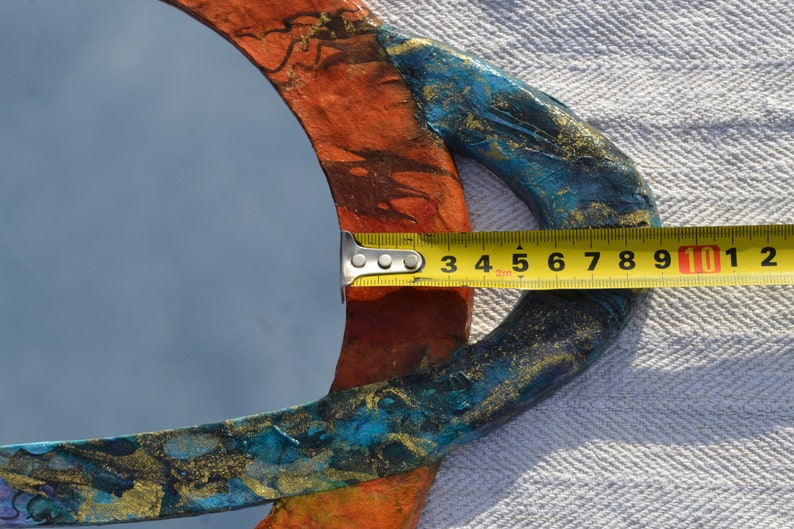 May include: A round mirror with a colorful, textured frame. The frame is painted with orange, blue, and gold accents. The mirror's diameter is approximately 4 inches as measured by a yellow tape measure.
