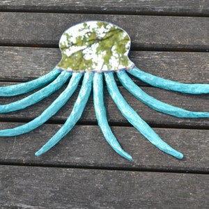 May include: A handmade mirror in the shape of an octopus. The mirror has a reflective surface and is surrounded by a turquoise-colored, textured frame with eight long, curved tentacles. The mirror is placed on a wooden surface.