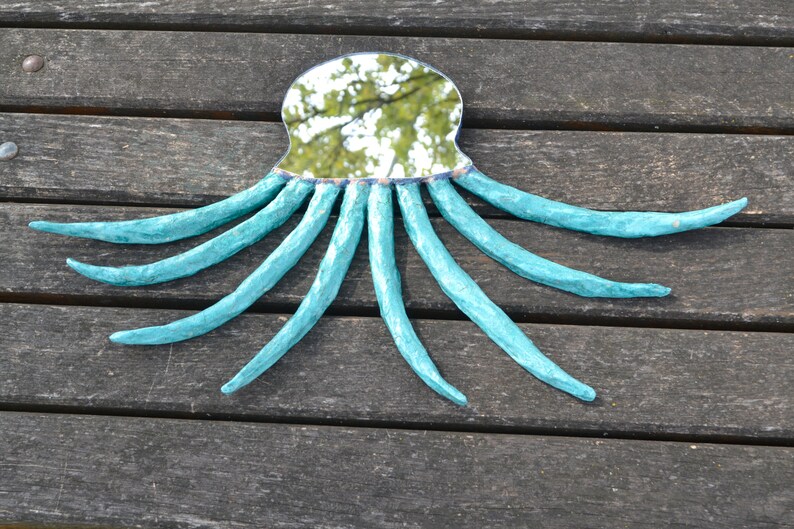 May include: A handmade mirror in the shape of an octopus. The mirror has a reflective surface and is surrounded by a turquoise-colored, textured frame with eight long, curved tentacles. The mirror is placed on a wooden surface.