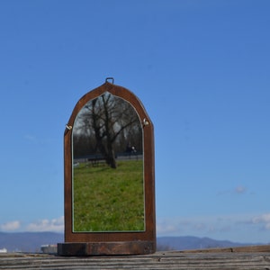 May include: A wooden framed mirror with an arched top, reflecting a tree and a bench in a grassy area. The mirror is sitting on a wooden base.