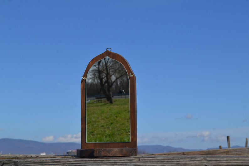 May include: A wooden framed mirror with an arched top, reflecting a tree and a bench in a grassy area. The mirror is sitting on a wooden base.