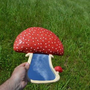 Pu&ograve; includere: Uno specchio a forma di fungo rosso e bianco con pois e un riflesso di cielo blu. Lo specchio &egrave; tenuto in una mano.