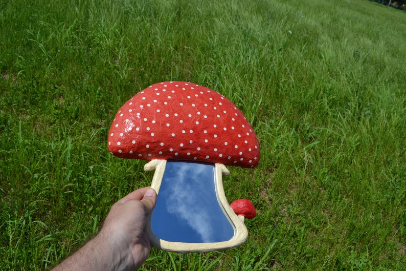 Pu&ograve; includere: Uno specchio a forma di fungo rosso e bianco con pois e un riflesso di cielo blu. Lo specchio &egrave; tenuto in una mano.