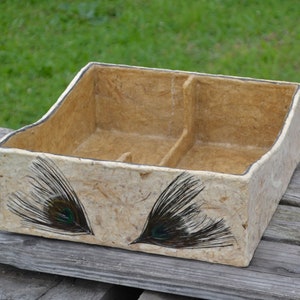 May include: A rectangular, handmade paper organizer with two compartments. The beige box features a dark trim and is decorated with two peacock feather designs in black and green. The box sits on a weathered wooden surface.