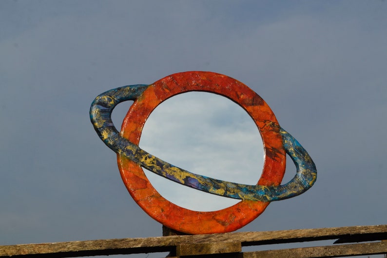 May include: A round mirror with a colorful, planet-like frame. The frame is painted with orange, blue, and gold hues. The mirror is mounted on a wooden structure against a cloudy sky.