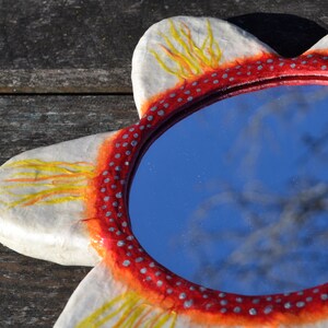 May include: A decorative, flower-shaped mirror with a red and white frame. The frame is adorned with small silver dots and yellow and orange accents. The mirror reflects a blue sky and tree branches.