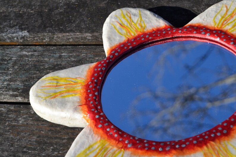 May include: A decorative, flower-shaped mirror with a red and white frame. The frame is adorned with small silver dots and yellow and orange accents. The mirror reflects a blue sky and tree branches.