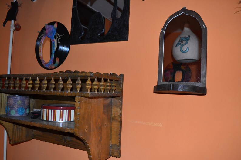 May include: A wooden shelf with a decorative carved trim and spindles. The shelf has two levels and is mounted on a wall. There are two items on the shelf, a round container with a red and white striped design and a small round container with a blue and white design. The wall is painted a light orange color.