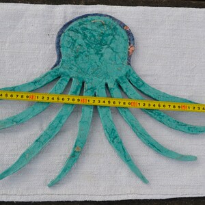 May include: A turquoise octopus-shaped wall hanging with a blue border, measured with a yellow tape measure. The octopus is approximately 24 inches wide. The tentacles are spread out, and the piece is on a white fabric surface.
