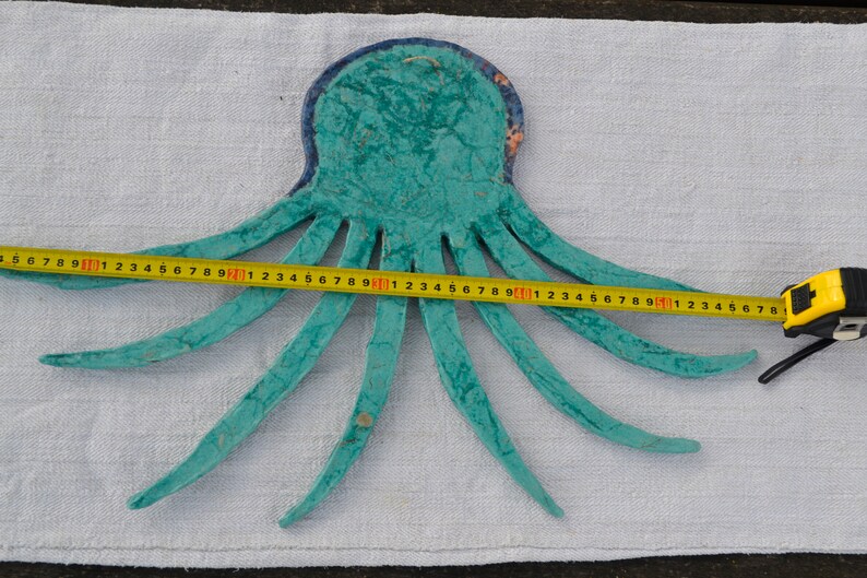 May include: A turquoise octopus-shaped wall hanging with a blue border, measured with a yellow tape measure. The octopus is approximately 24 inches wide. The tentacles are spread out, and the piece is on a white fabric surface.