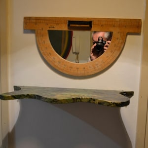 May include: A wall-mounted mirror shaped like a large wooden protractor with black numerical markings and a central black handle, positioned above a floating shelf with a mottled green and yellow textured surface finish.