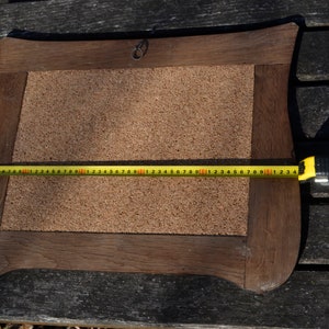 May include: A brown wooden frame with a corkboard insert. The frame has a decorative, slightly curved shape. A yellow measuring tape is placed across the corkboard, measuring 50 inches.