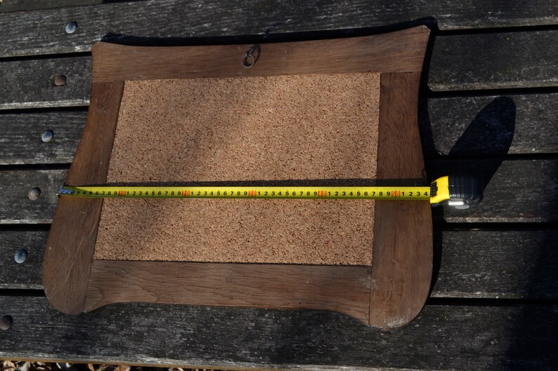 May include: A brown wooden frame with a corkboard insert. The frame has a decorative, slightly curved shape. A yellow measuring tape is placed across the corkboard, measuring 50 inches.