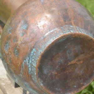May include: A large copper pitcher with a worn, aged patina. The bottom of the pitcher has a circular design with a green patina.