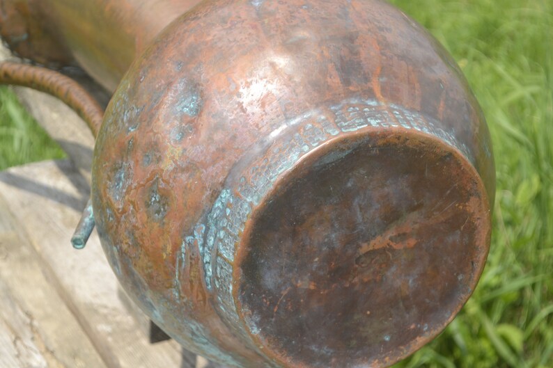 May include: A large copper pitcher with a worn, aged patina. The bottom of the pitcher has a circular design with a green patina.