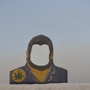 Puede incluir: Un recorte de cartón de la cabeza de una persona con una hoja de marihuana verde en el hombro. El recorte está pintado en tonos de azul, amarillo y marrón.