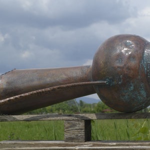 May include: A large, copper, hammered pitcher with a long, curved spout and a handle. The pitcher is resting on a wooden fence rail with a green field in the background.