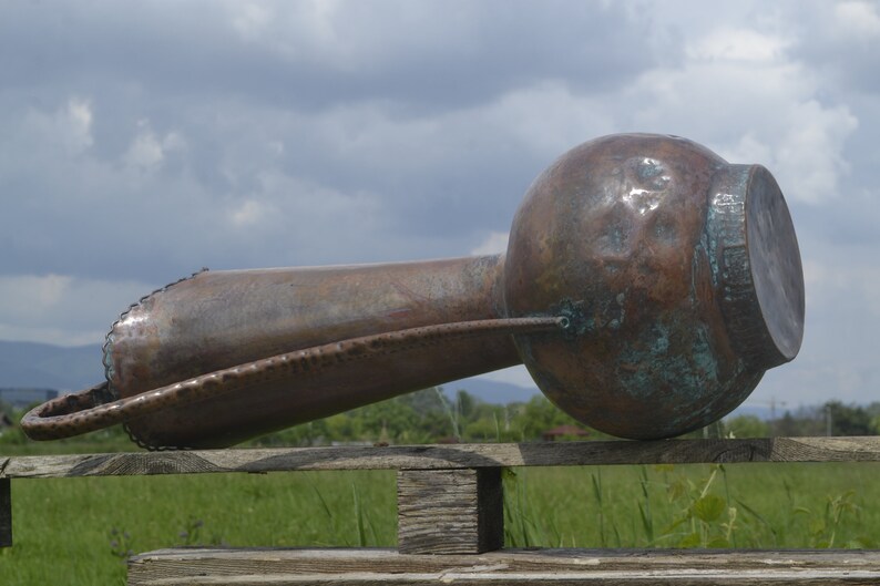 May include: A large, copper, hammered pitcher with a long, curved spout and a handle. The pitcher is resting on a wooden fence rail with a green field in the background.