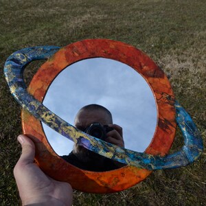 May include: A round mirror with a colorful, planet-like frame. The frame is primarily orange and blue with gold accents. The mirror reflects the sky and a person holding a camera.