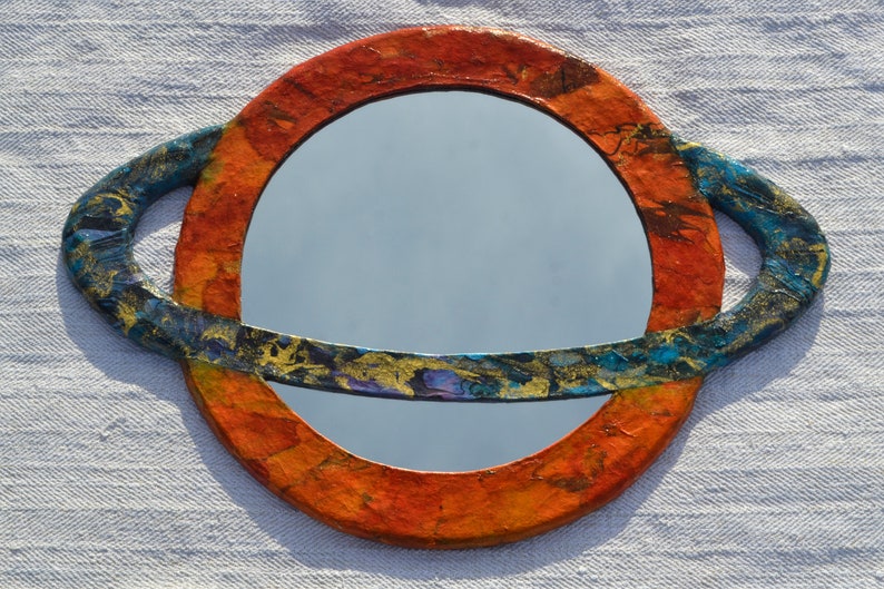 May include: A round mirror with a decorative frame resembling a planet. The frame is primarily orange with blue and gold accents. The mirror's surface reflects the sky. The overall design is artistic and unique.