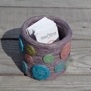 May include: A purple, cylindrical, handmade container with colorful swirls and dots. The container is filled with white business cards that say "Unique Memories" and "Holders - Holland, MI".