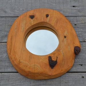 Puede incluir: Un espejo redondo de madera con un acabado de madera natural. El espejo est&aacute; encajado en un marco de madera grueso con una textura rugosa y org&aacute;nica. El marco tiene varios nudos e imperfecciones, lo que le da un aspecto r&uacute;stico.