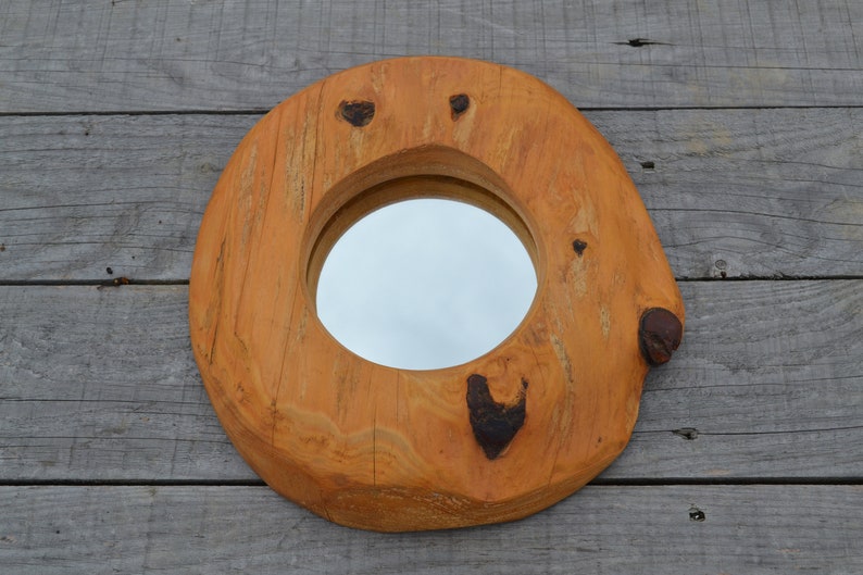 Puede incluir: Un espejo redondo de madera con un acabado de madera natural. El espejo est&aacute; encajado en un marco de madera grueso con una textura rugosa y org&aacute;nica. El marco tiene varios nudos e imperfecciones, lo que le da un aspecto r&uacute;stico.