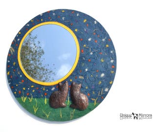 Op de afbeelding: Een ronde, blauwe spiegel met een gele lijst en een strooiing van kleurrijke bloemen. Twee bruine katten zijn aan de onderkant van de spiegel gebeeldhouwd en kijken naar de reflectie.