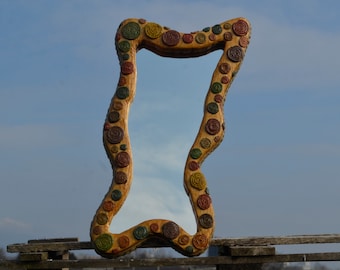 May include: A large, wavy-edged mirror with a wooden frame. The frame is decorated with colorful, circular, raised designs in shades of brown, green, yellow, and red. The mirror is outdoors against a blue sky.