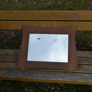 May include: A wooden framed mirror with a slightly curved top and bottom edge, reflecting a cloudy sky with a small bird flying in the distance.