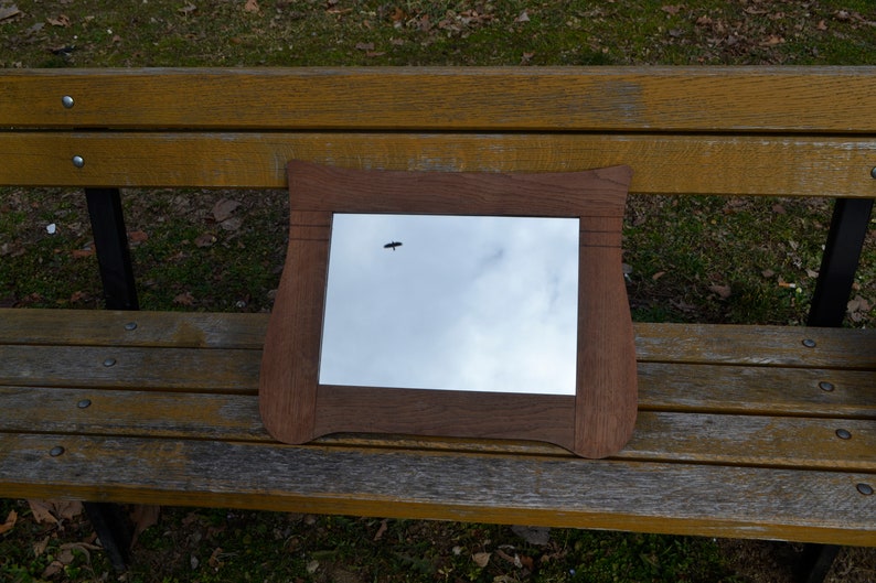 May include: A wooden framed mirror with a slightly curved top and bottom edge, reflecting a cloudy sky with a small bird flying in the distance.