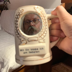May include: A cream-colored ceramic beer stein featuring a circular mirror on the front, surrounded by a gold-colored laurel wreath design. Below the mirror is a rectangular plaque with the black text, Mit Dir trinke ich am liebsten.