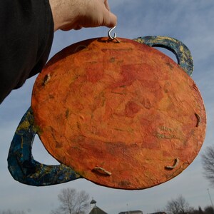 May include: A handmade planet decoration with a textured orange surface and blue rings. The planet is held by a silver hook. The background shows a blue sky and a few trees.