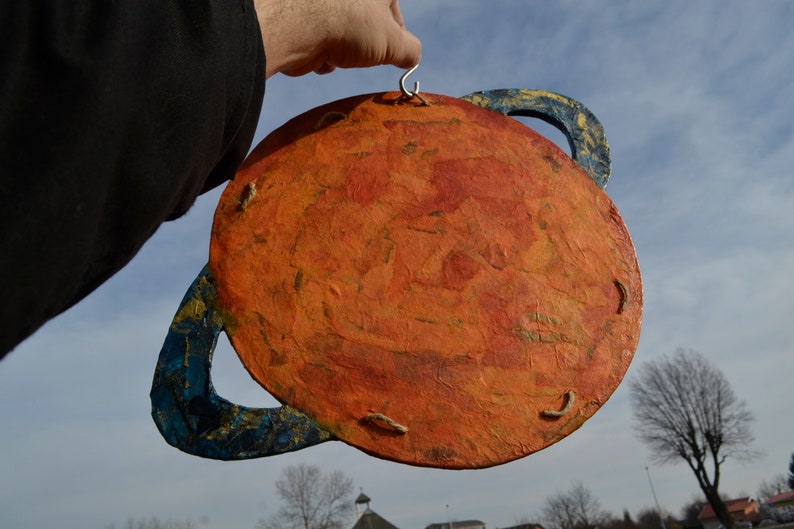 May include: A handmade planet decoration with a textured orange surface and blue rings. The planet is held by a silver hook. The background shows a blue sky and a few trees.