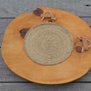 Puede incluir: Una bandeja redonda de madera con un acabado de madera natural. La bandeja tiene un c&iacute;rculo de cuerda en el centro y dos asas de corcho a los lados.