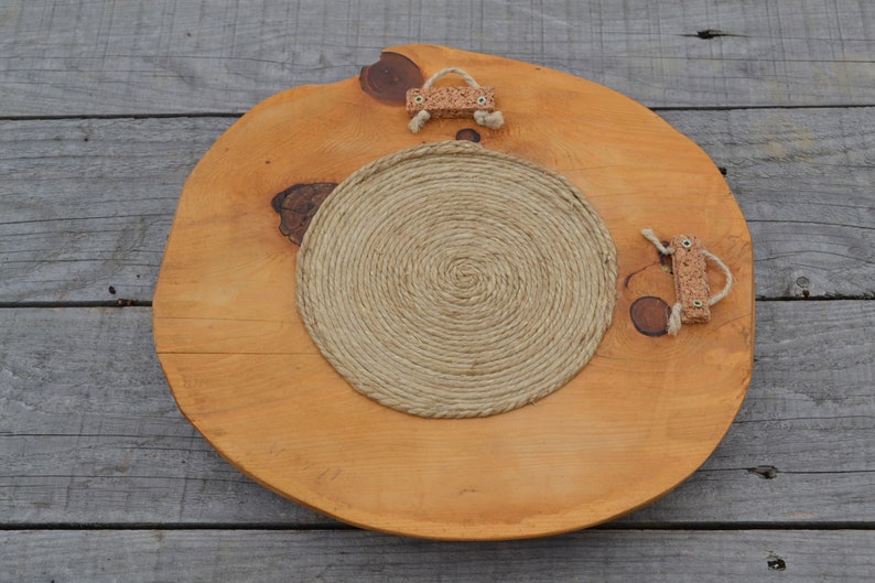 Puede incluir: Una bandeja redonda de madera con un acabado de madera natural. La bandeja tiene un c&iacute;rculo de cuerda en el centro y dos asas de corcho a los lados.